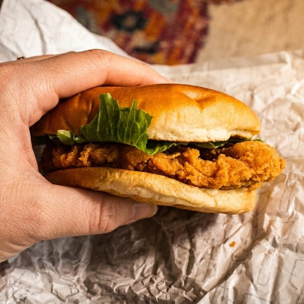 Hand holding a crispy fried chicken sandwich with lettuce on a bun, wrapped in paper.